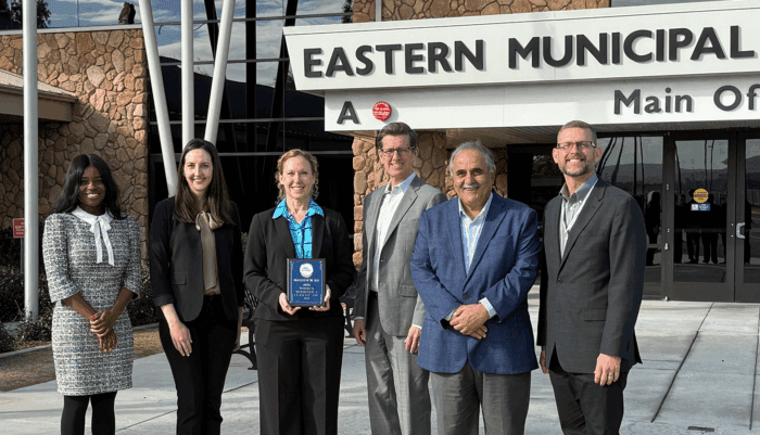 Three women and three men stand in front of the entrance to Eastern Municipal Water District's main office. The woman in the middle is holding a blue plaque, which names Woodard & Curran Consultant of the Year.