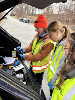 A man in a safety vest and blaze orange hat demonstrates to two women, also wearing safety vests, how to collect field data on a mobile phone during an Illicit Discharge Detection and Elimination (IDDE) investigation. 