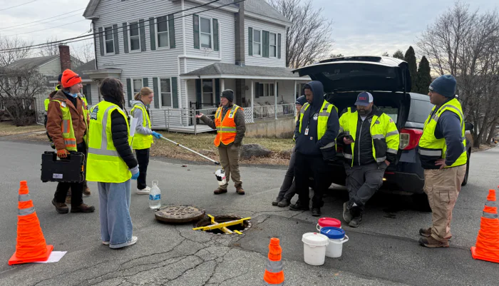 A group of seven people stand around a man hole in front of a residential hole learning how to conduct field investigations of stormwater infrastructure for an have Illicit Discharge Detection and Elimination (IDDE) program.