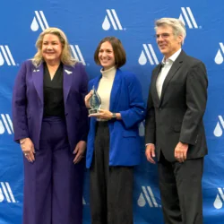Stephanie Estabrook, pictured center, is dressed in a cream turtleneck with blue blazer and dark gray/black pinstriped slacks, holding a glass water drop trophy. The woman to the left and man to the right are both dressed in business suits. The three are posed in front of a step and repeat blue backdrop with the white AWWA logo.